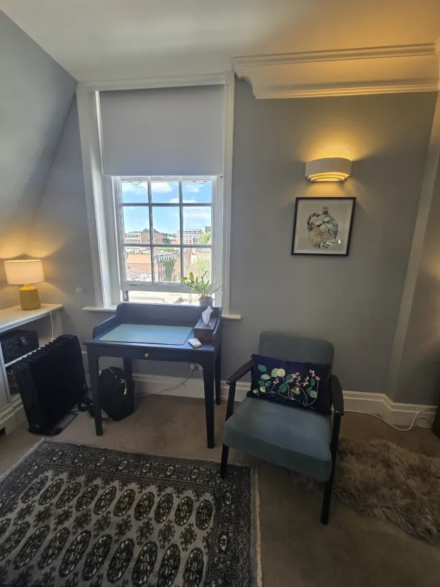 Therapy room at Eaton Gate Practice with a desk, armchair, rug, and city-facing window